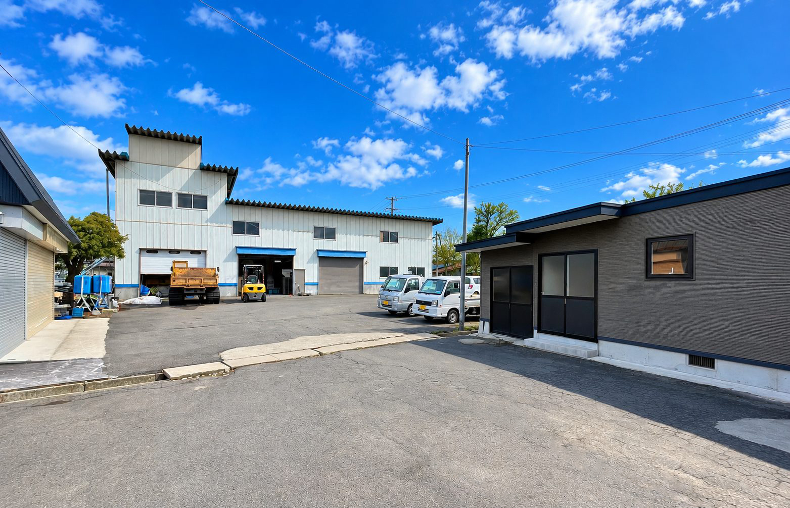 作業場の建屋とフォークリフト、ダンプトラック、軽トラックが並ぶ事業所の敷地。右手に事務所棟